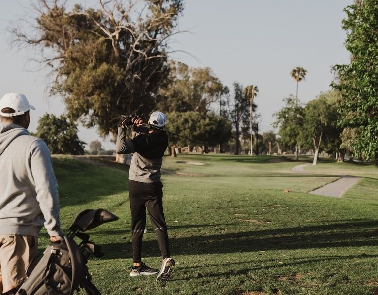Golfer practicing a shot on a tree-lined course beside a golf bag